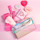 A glitter-themed gift bundle arranged on a pink background, featuring a metallic zip pouch, pink and white plush accessories, a heart-shaped compact, lip product tubes, and a small boxed item. Text across the lower portion reads, '5 brand-new Glitter Girl products together in one exclusive Gift Bundle.