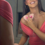 A woman with long dark hair smiles while looking into a mirror, wearing a bright coral pink off-the-shoulder top. Makeup bags and cosmetics are visible on the counter in front of her, with vanity lights glowing in the background.