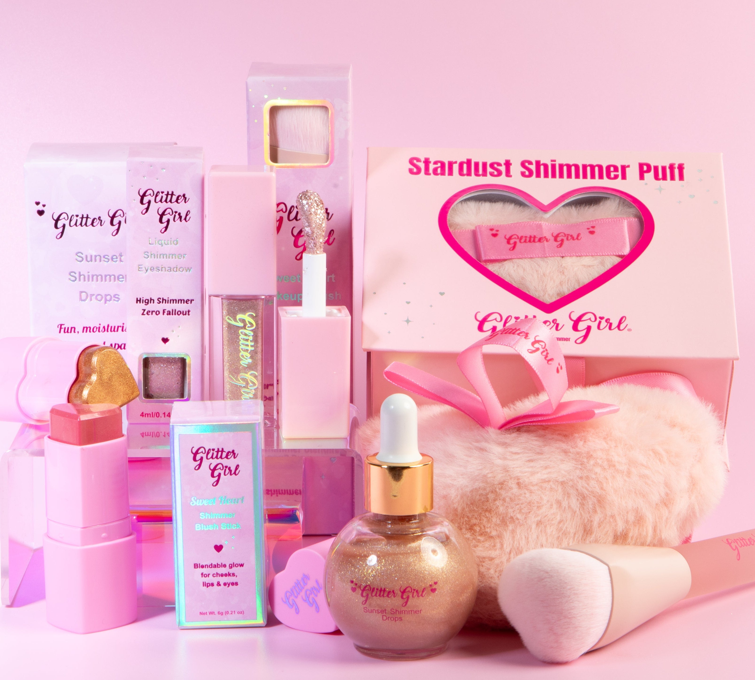 A beauty gift set from Glitter Girl featuring pink and gold cosmetic products including shimmer drops in a bottle, lip gloss, face shimmer puff and a fluffy pink brush, arranged in front of branded packaging boxes on a solid pink background.