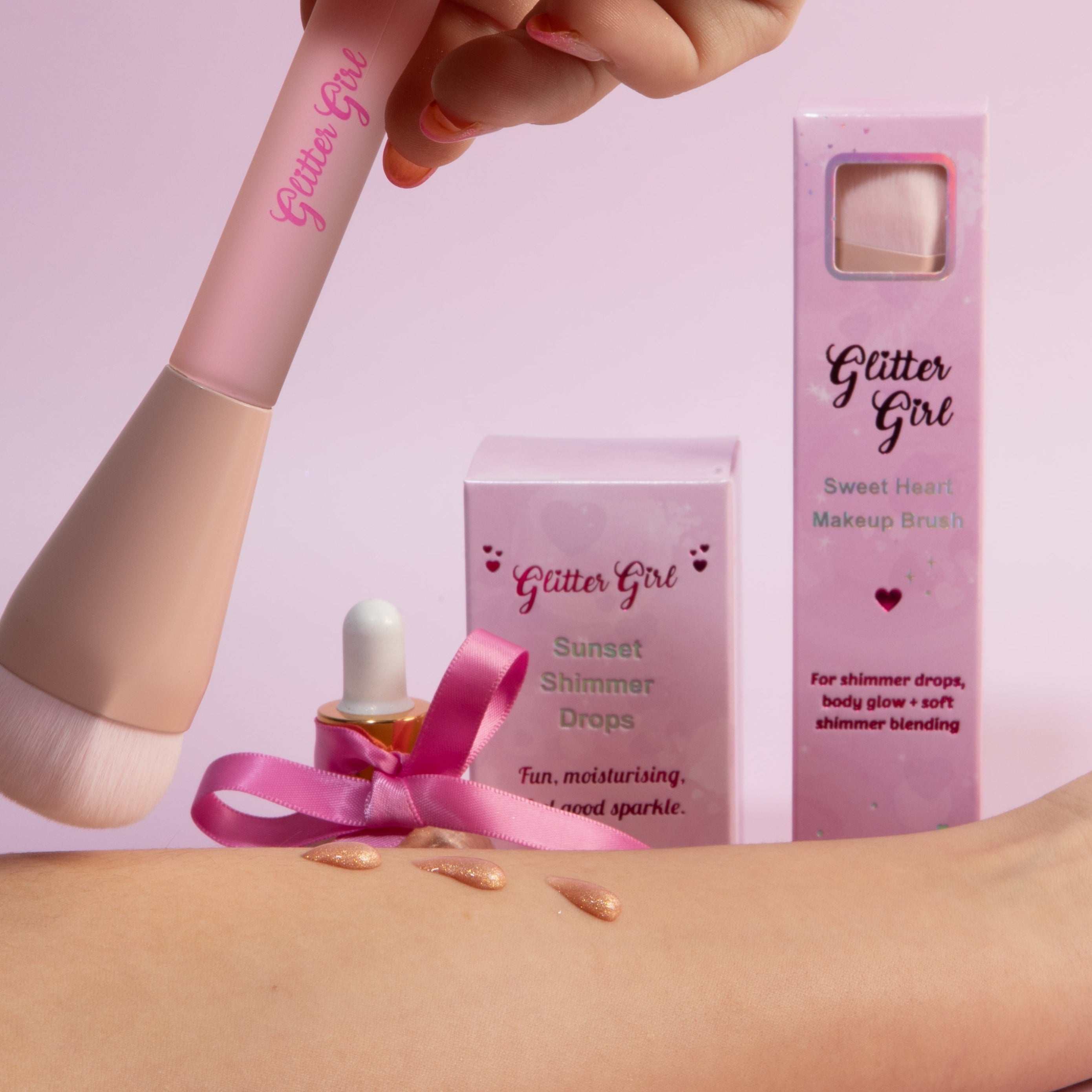 A beauty product set from Glitter Girl, including a pink makeup brush, a pink applicator bottle with a pink ribbon, a pink box, and a rose gold glitter makeup dropper bottle held in a hand, all set against a solid pink background.