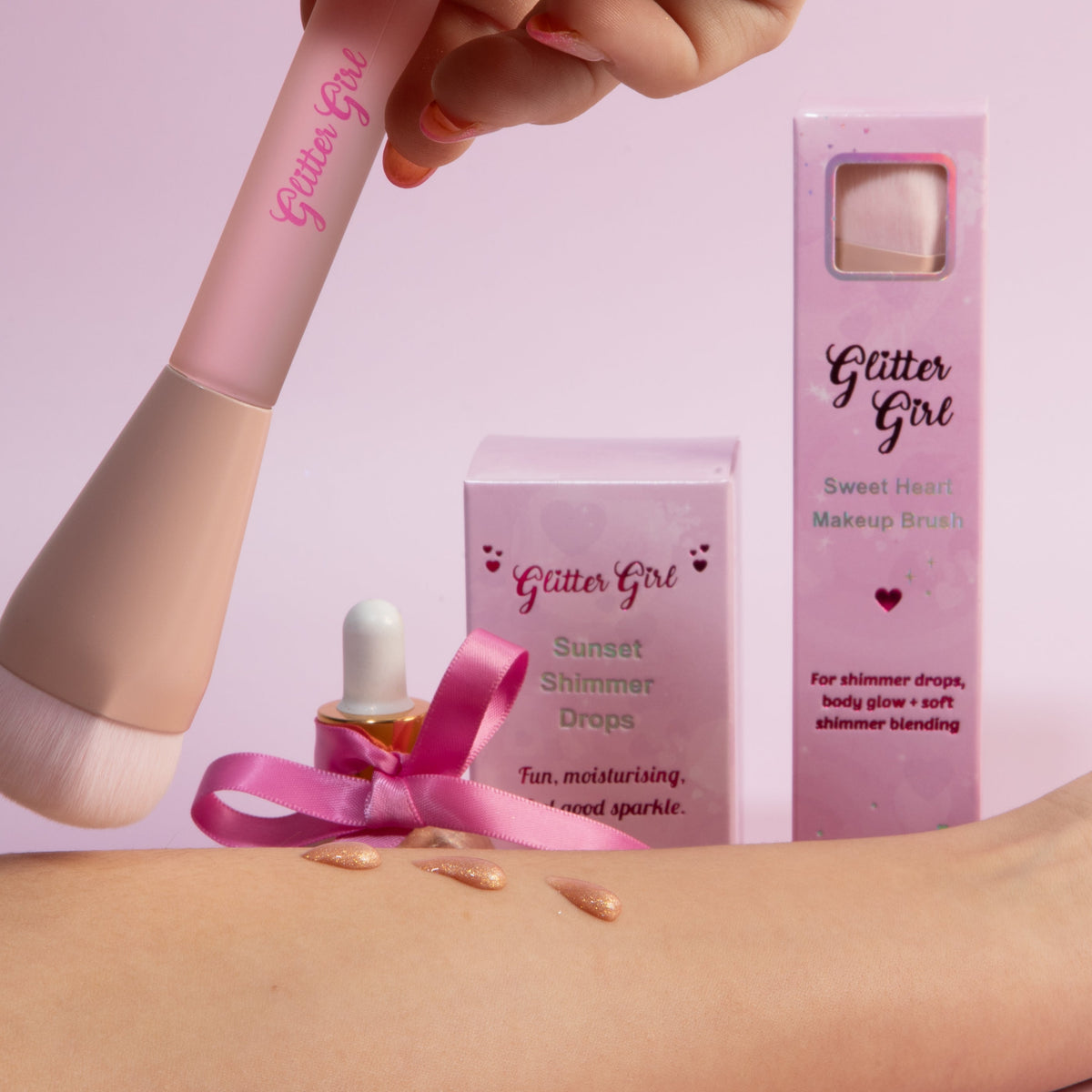 A beauty product set from Glitter Girl, including a pink makeup brush, a pink applicator bottle with a pink ribbon, a pink box, and a rose gold glitter makeup dropper bottle held in a hand, all set against a solid pink background.